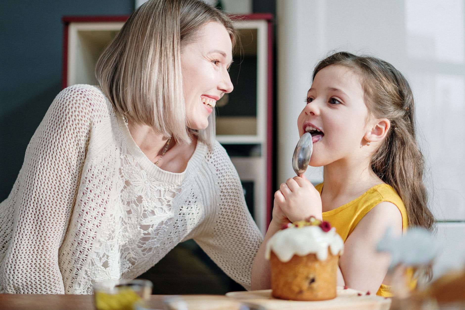 woman in white knit sweater smiling while little girl licking icing on her spoon