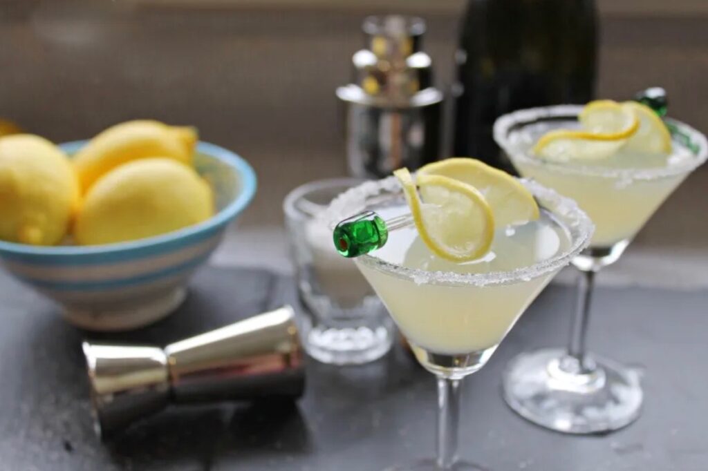 Two martini glasses filled with a yellow coloured cocktail. They are topped with twists of lemon. Behind is a bowl of lemons and some cocktail shakers