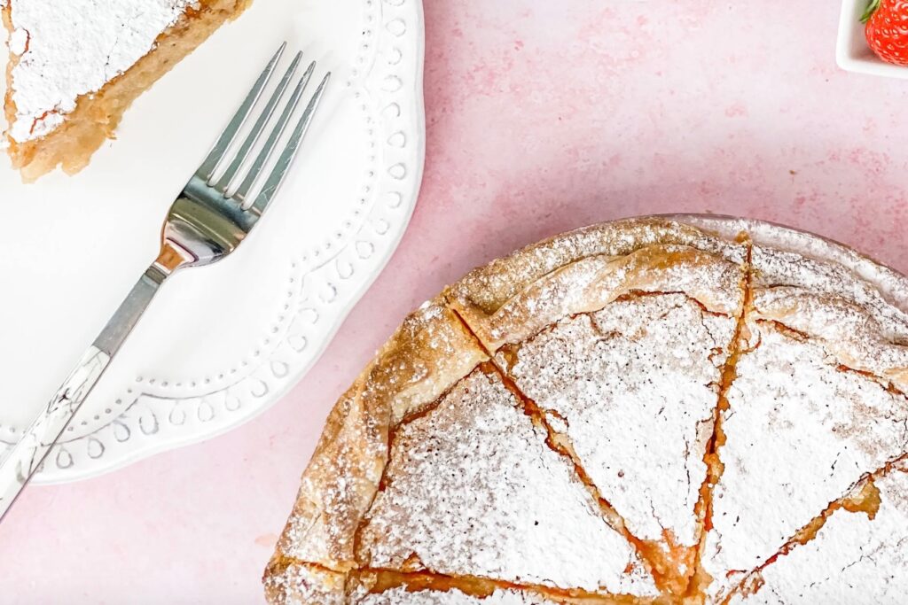 A pie topped with icing sugar next to a white plate with a fork on it