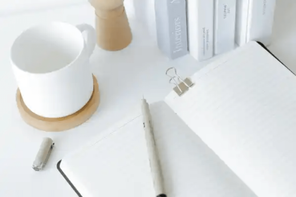 A notepad open on a blank page with a white pen lying across it. They are on a white desk with a white mug 