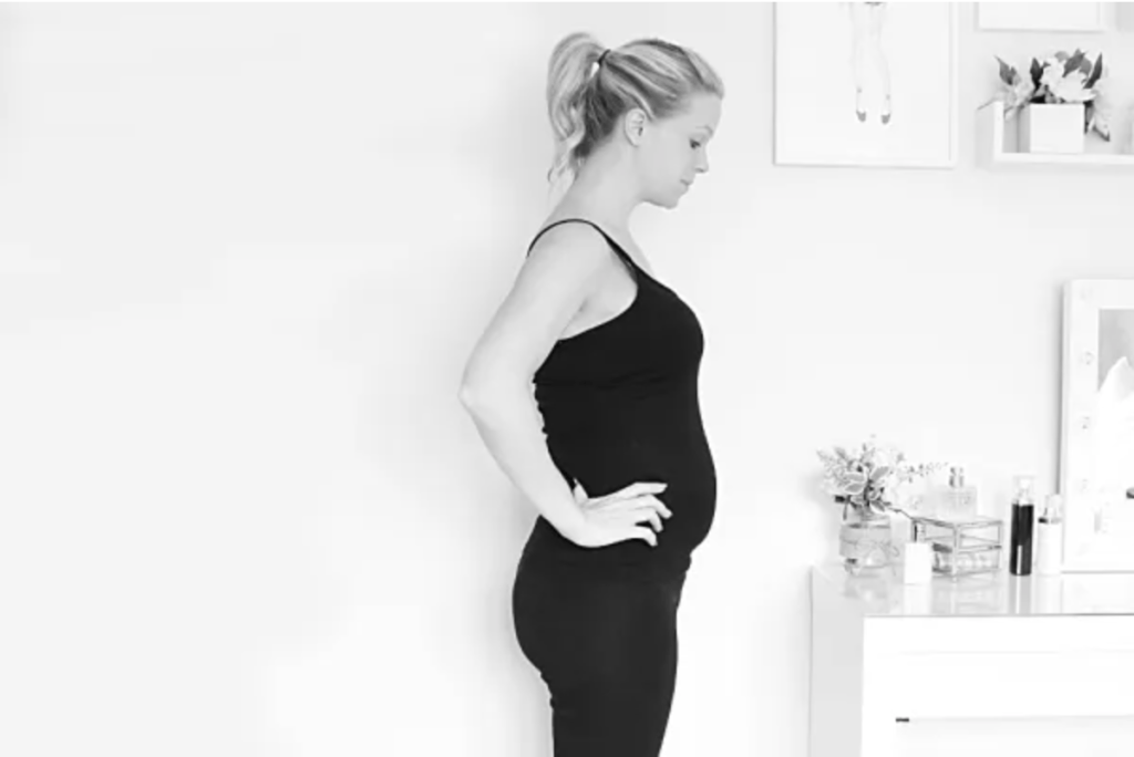 A black and white picture of a woman that is 19 weeks pregnant. She is standing side on to the camera in an all black outfit with her hands on her hips.