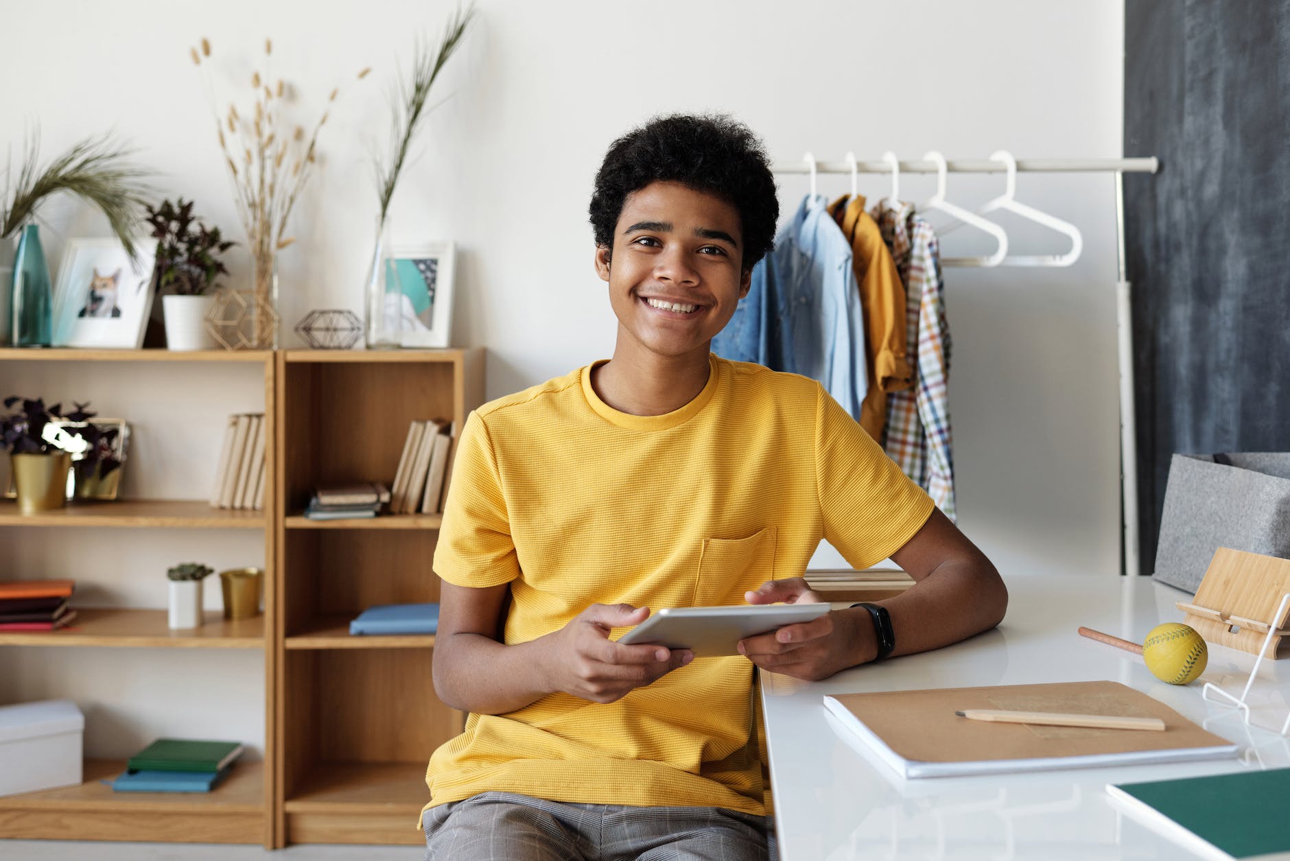 boy in yellow crew neck t shirt sitting on chair. He has dark skin and hair and is smiling at the camera while holding an iPad