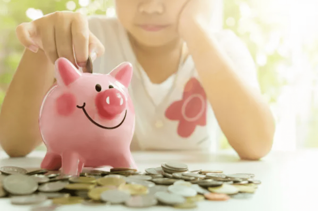 A child sitting at a table with a pink piggy bank. They are dropping a coin into the back