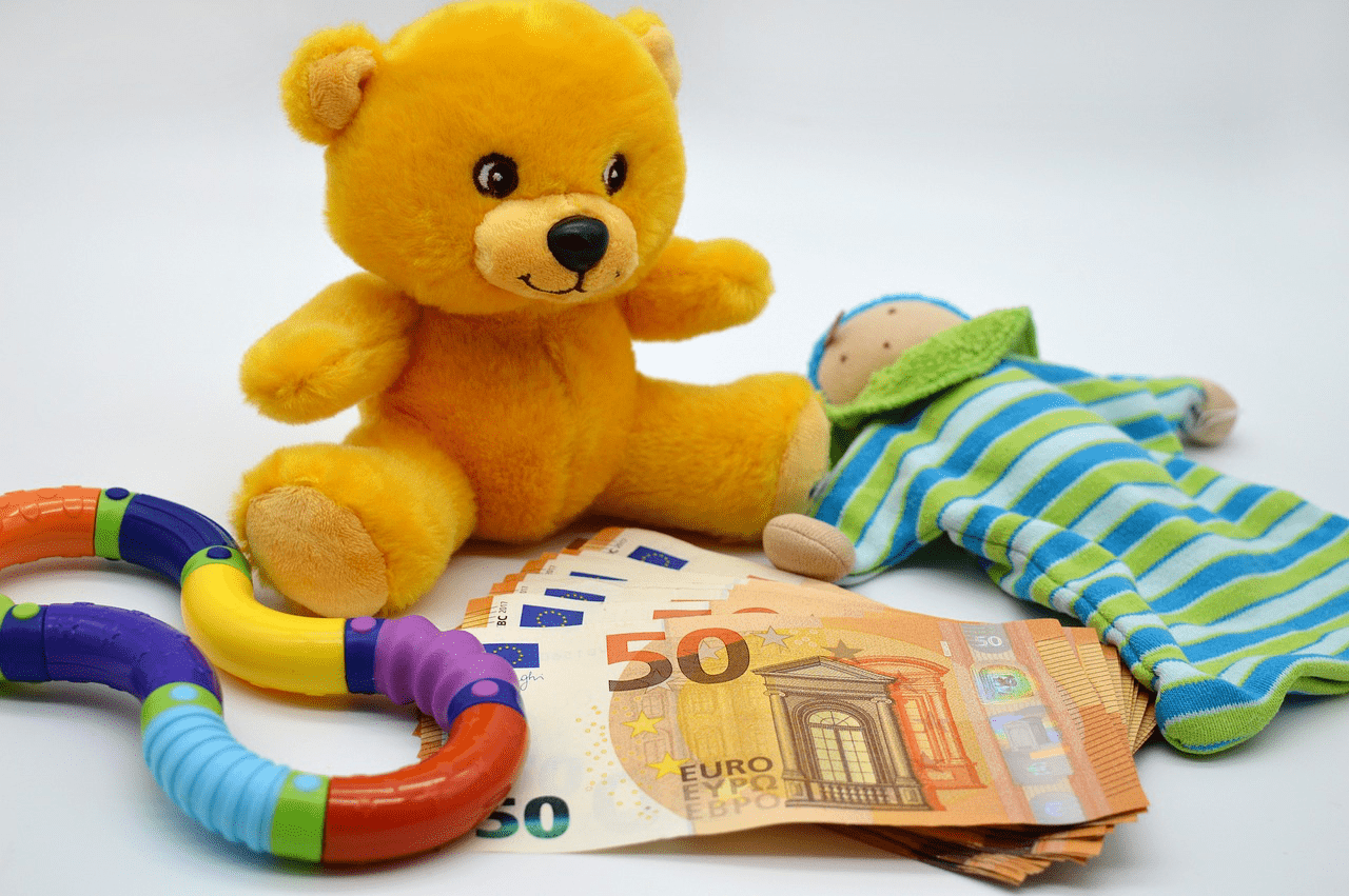 A pile of 50 Euro notes is in the centre of the image. A small yellow teddybear, a little doll and a plastic teething toy are next to it.