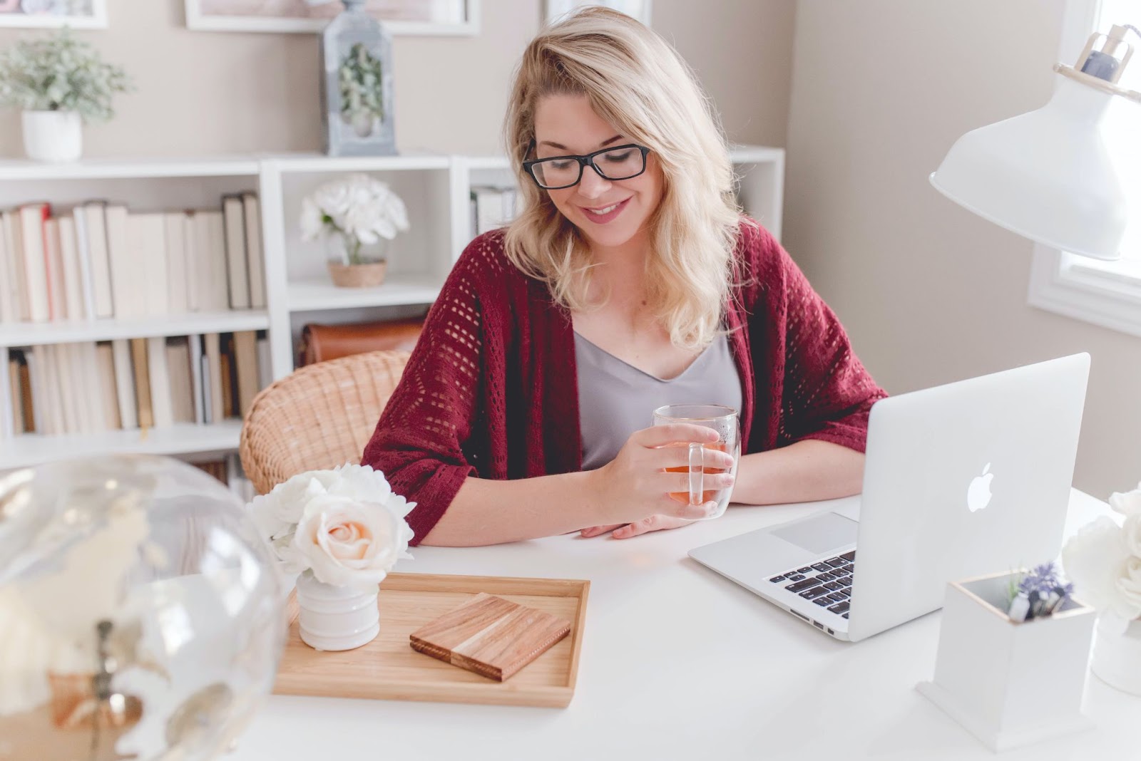 A woman with blonde hair and glasses sitting at a desk. She has a mug in her hand and is looking down at a small floral decoration. A Mac laptop is open in front of her, and in the background is a white bookcase.