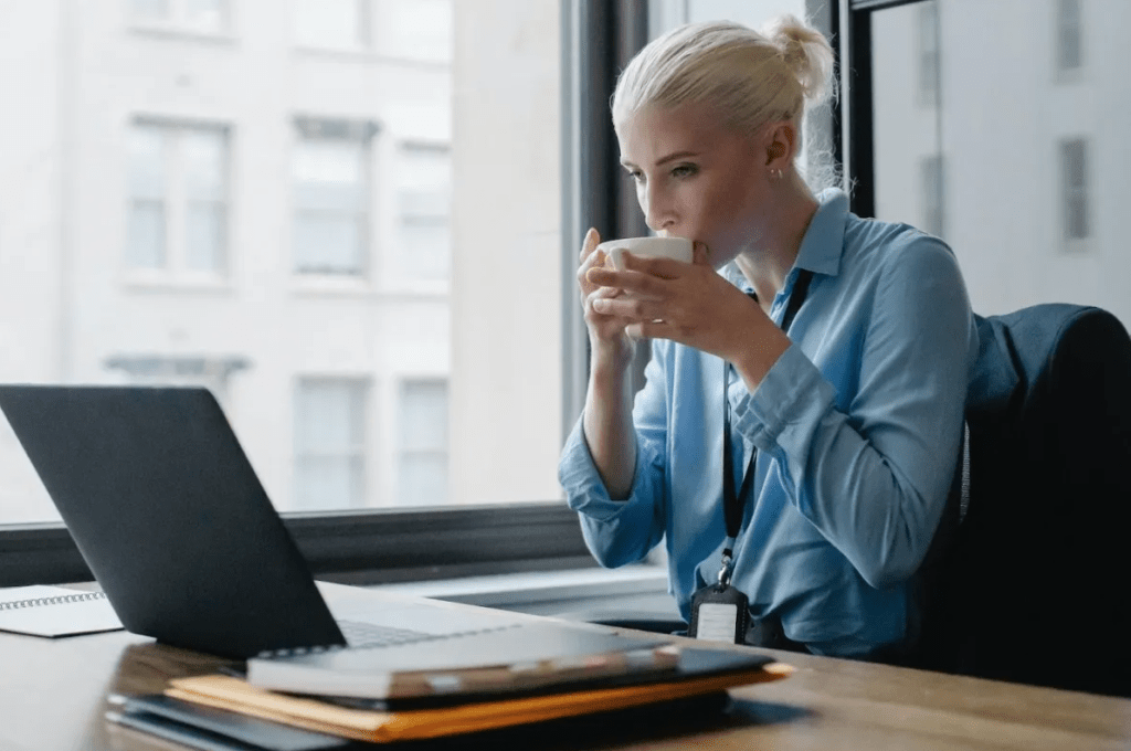 A woman sitting at a desk and taking a sip from a mug while she looks at her computer screen.