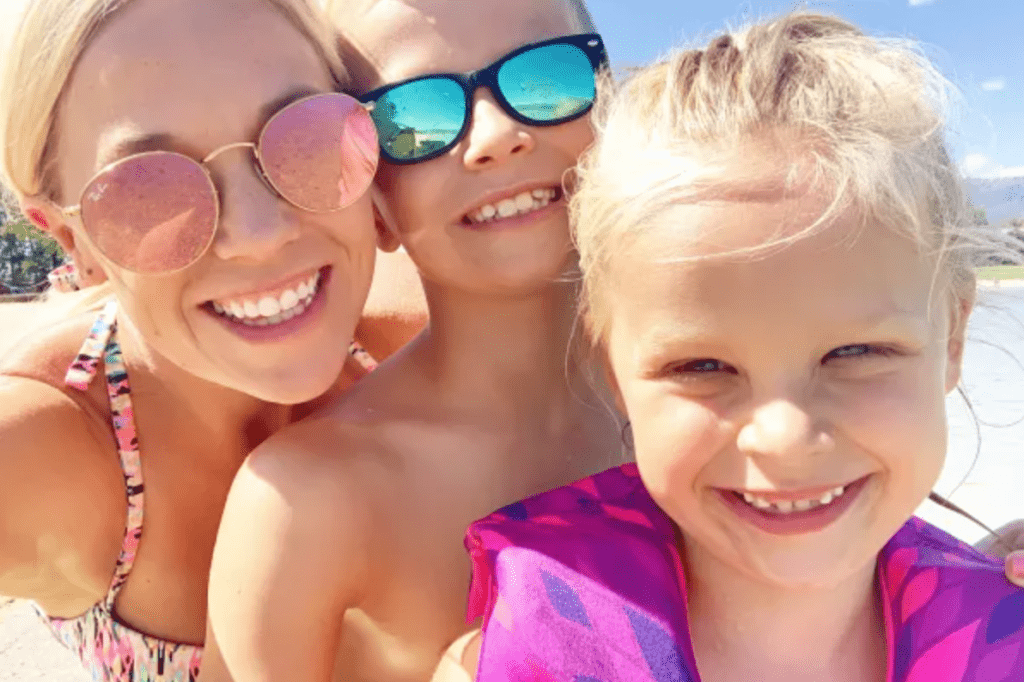 A mum, son and daughter up close to the camera for a selfie. They are wearing sunglasses and swimwear and smiling in the sunshine