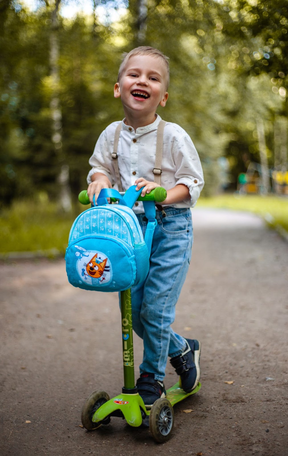 A young boy standing on a scooter on a bath surrounded by trees. He smiling and clearly enjoying himself.