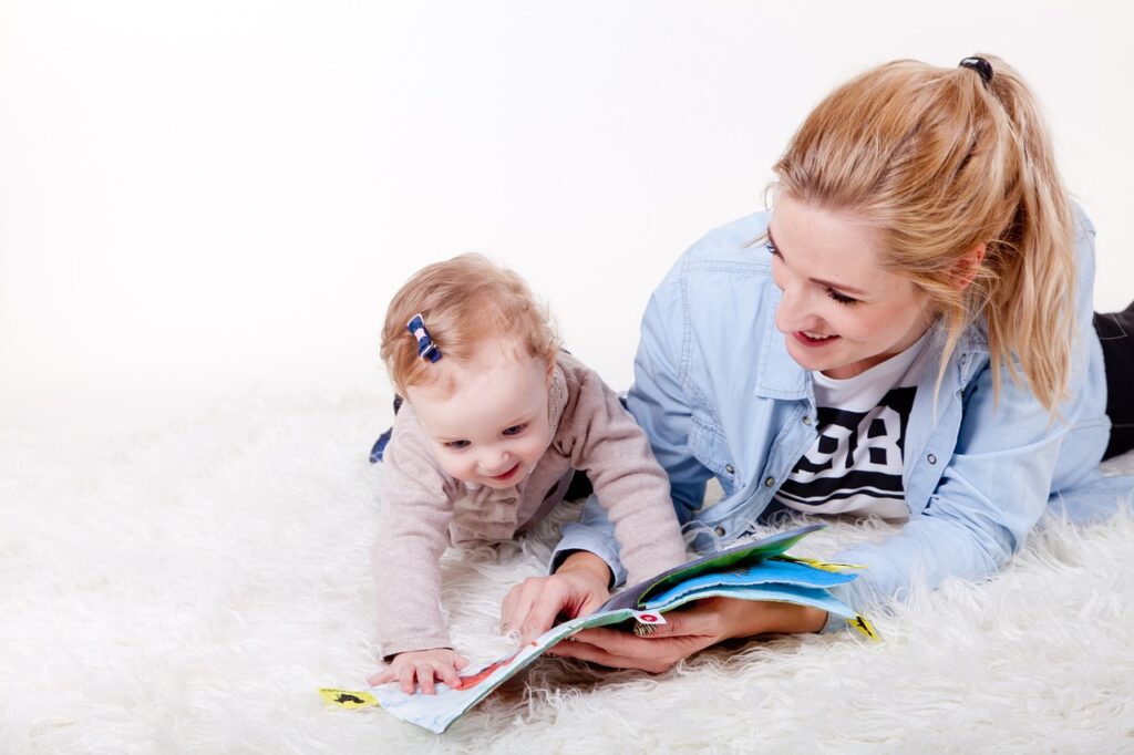 A woman is reading a book to her baby on a white rug.