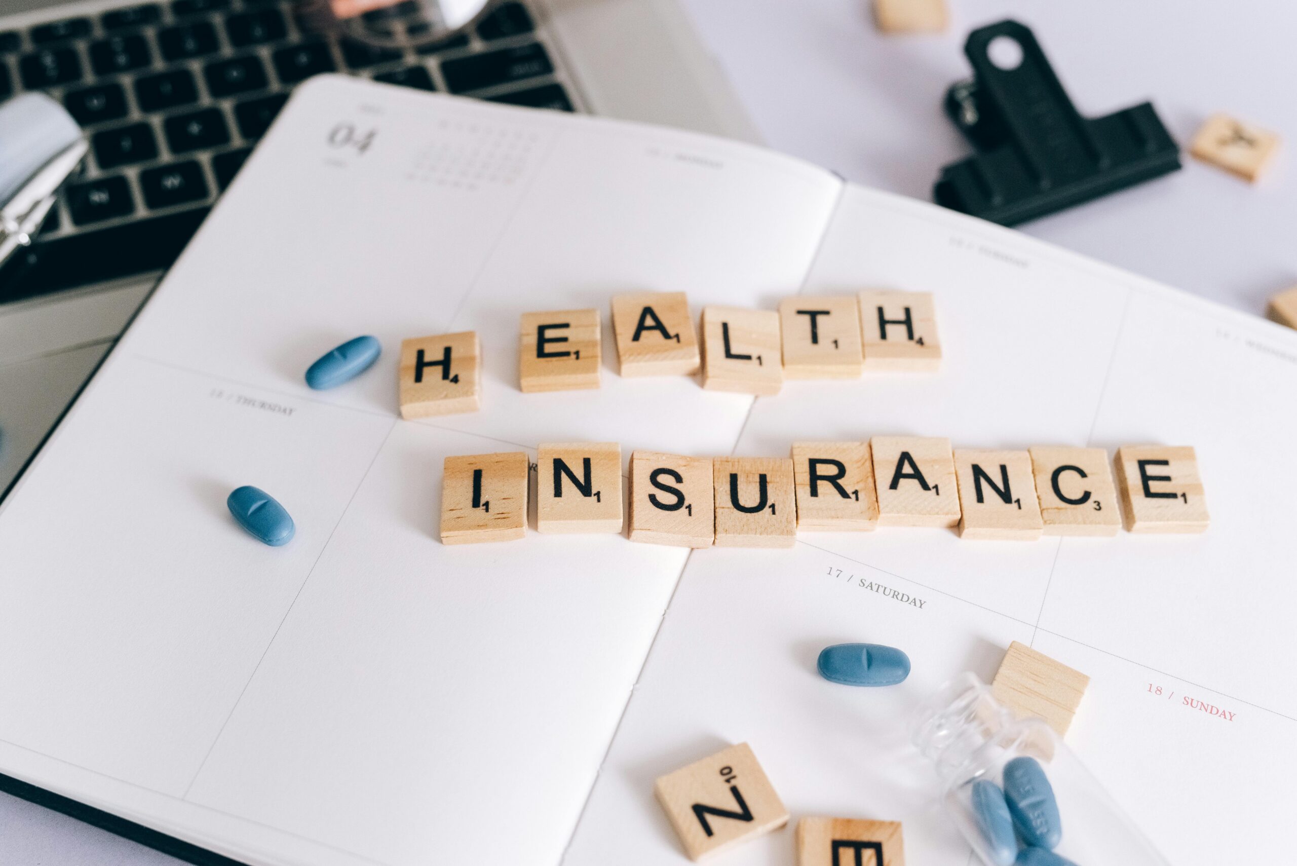 Wooden letter tiles spell out "expats health insurance" across an open planner, with pills and a keyboard nearby.